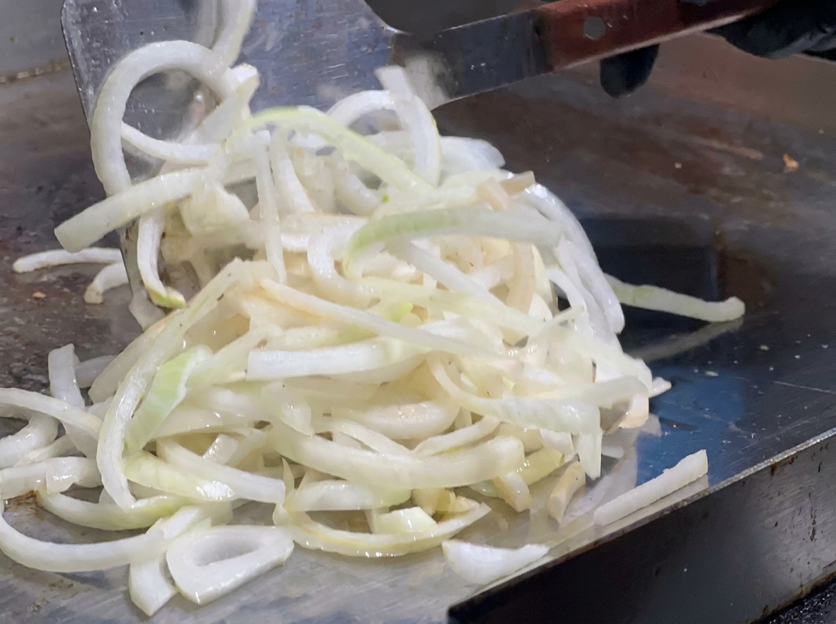 Zwiebelstreifen werden mit einem großen Metallspatel auf dem heißen Grill verteilt.