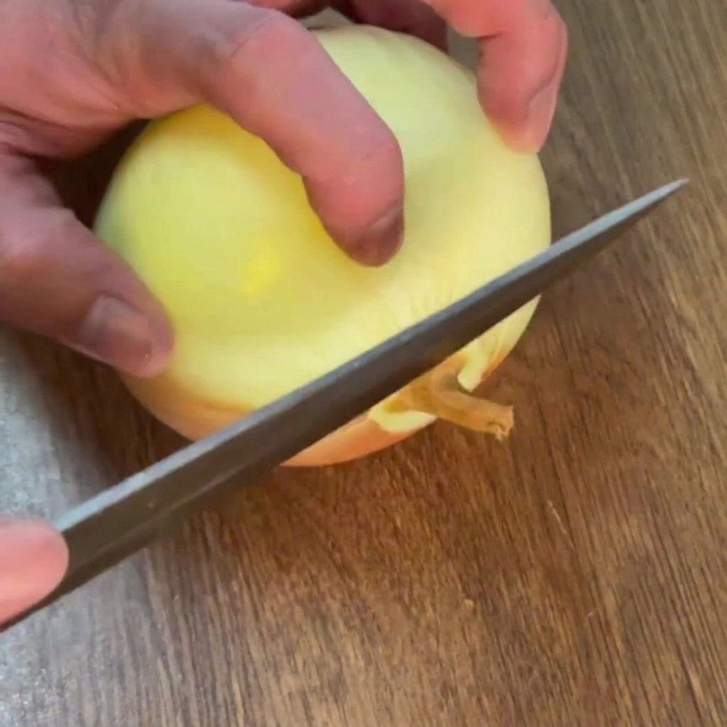 Hände schneiden mit einem Messer das Wurzelende einer geschälten gelben Zwiebel auf einem Holzschneidebrett ab.
