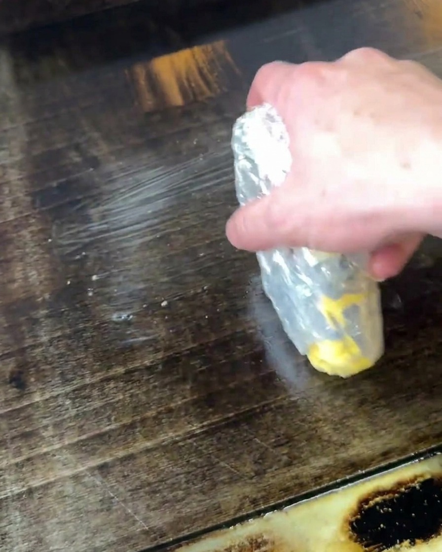 Eine Hand hält ein Stück Butter und reibt es direkt auf eine heiße, dunkle Grillplatte, um sie zu schmelzen.