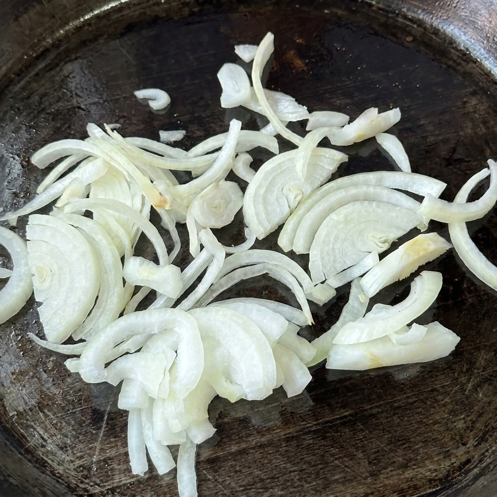Ein Haufen roher, in dünne Scheiben geschnittener weißer Zwiebeln auf einer eingefetteten, dunklen Grillplatte.