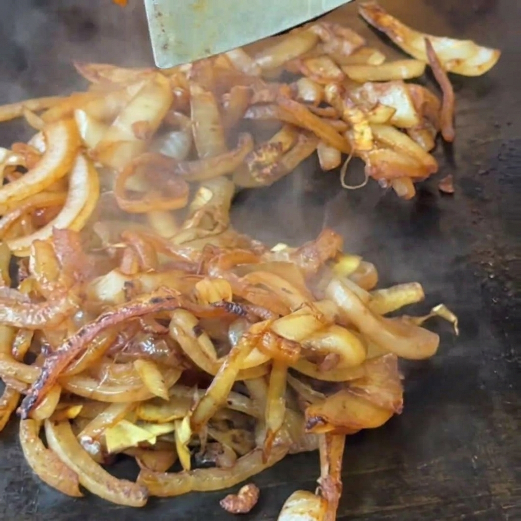 Ein Pfannenwender rührt weich werdende, leicht gebräunte Zwiebelscheiben auf einer heißen Grillplatte um.