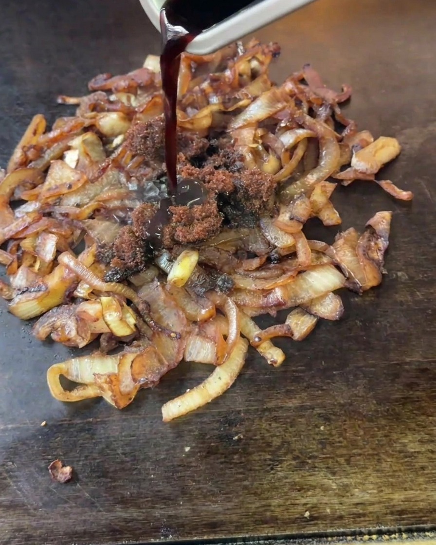 Dunkler Essig und brauner Zucker werden aus einem kleinen Metallbehälter über die kochenden Zwiebeln gegossen.