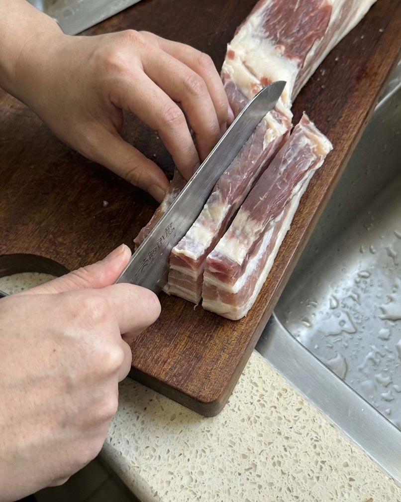 Roher Schweinebauch wird mit einem scharfen Küchenmesser in lange, dicke Streifen geschnitten.