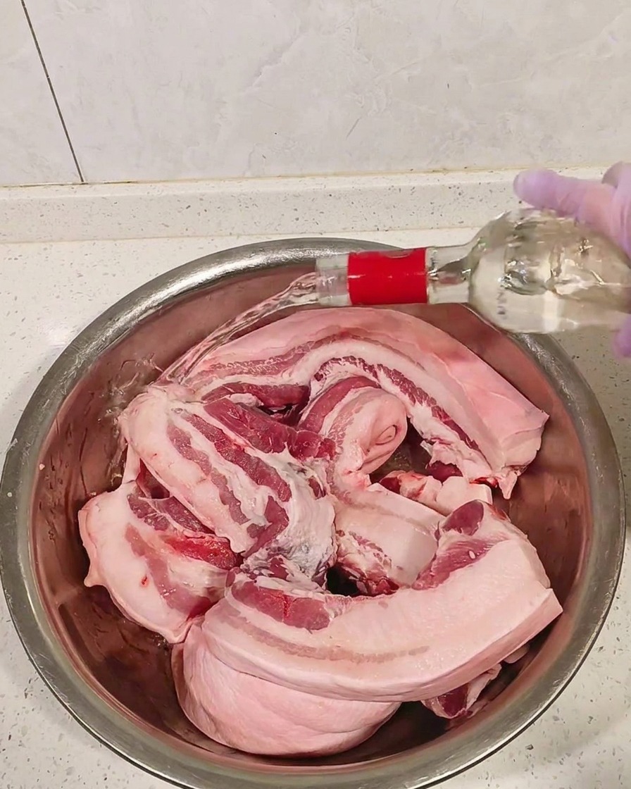 Rohe Schweinebauchstücke in einer Metallschüssel, übergossen mit klarem weißen Schnaps.