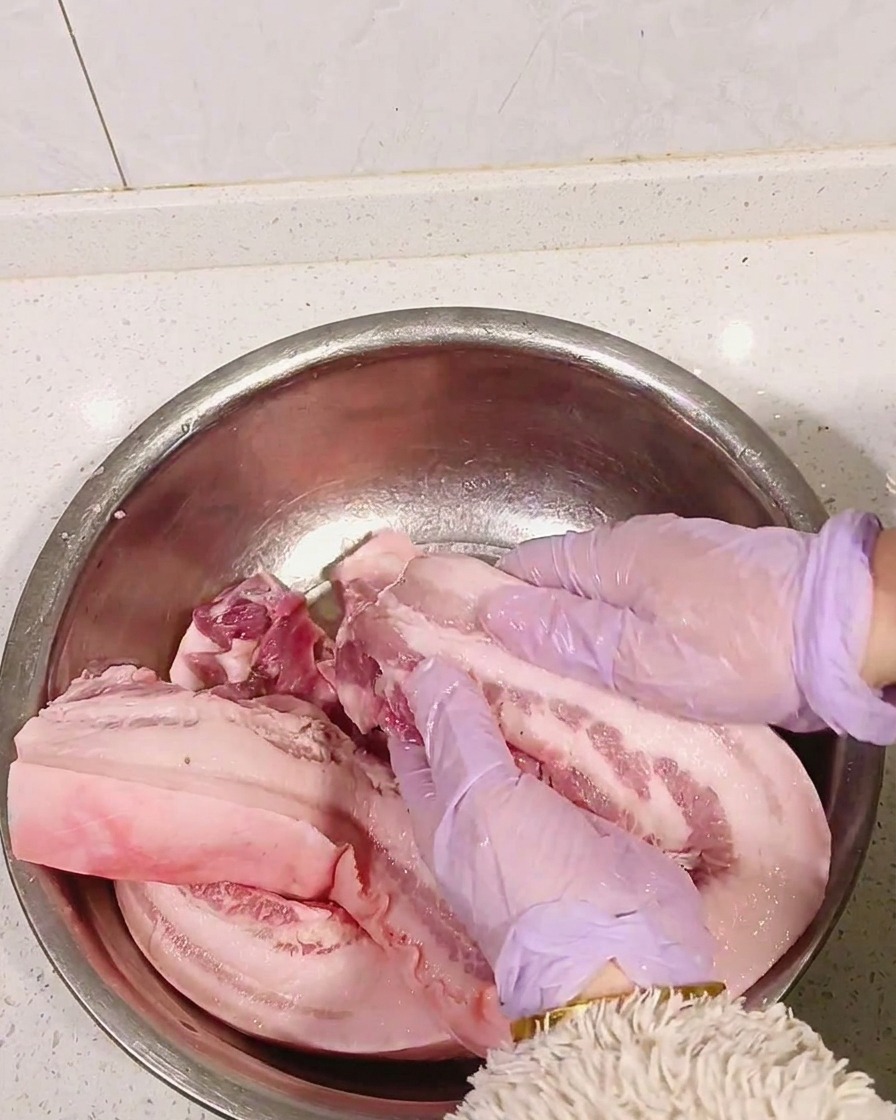Hände in lila Handschuhen massieren weißen Schnaps in den rohen Schweinebauch ein.