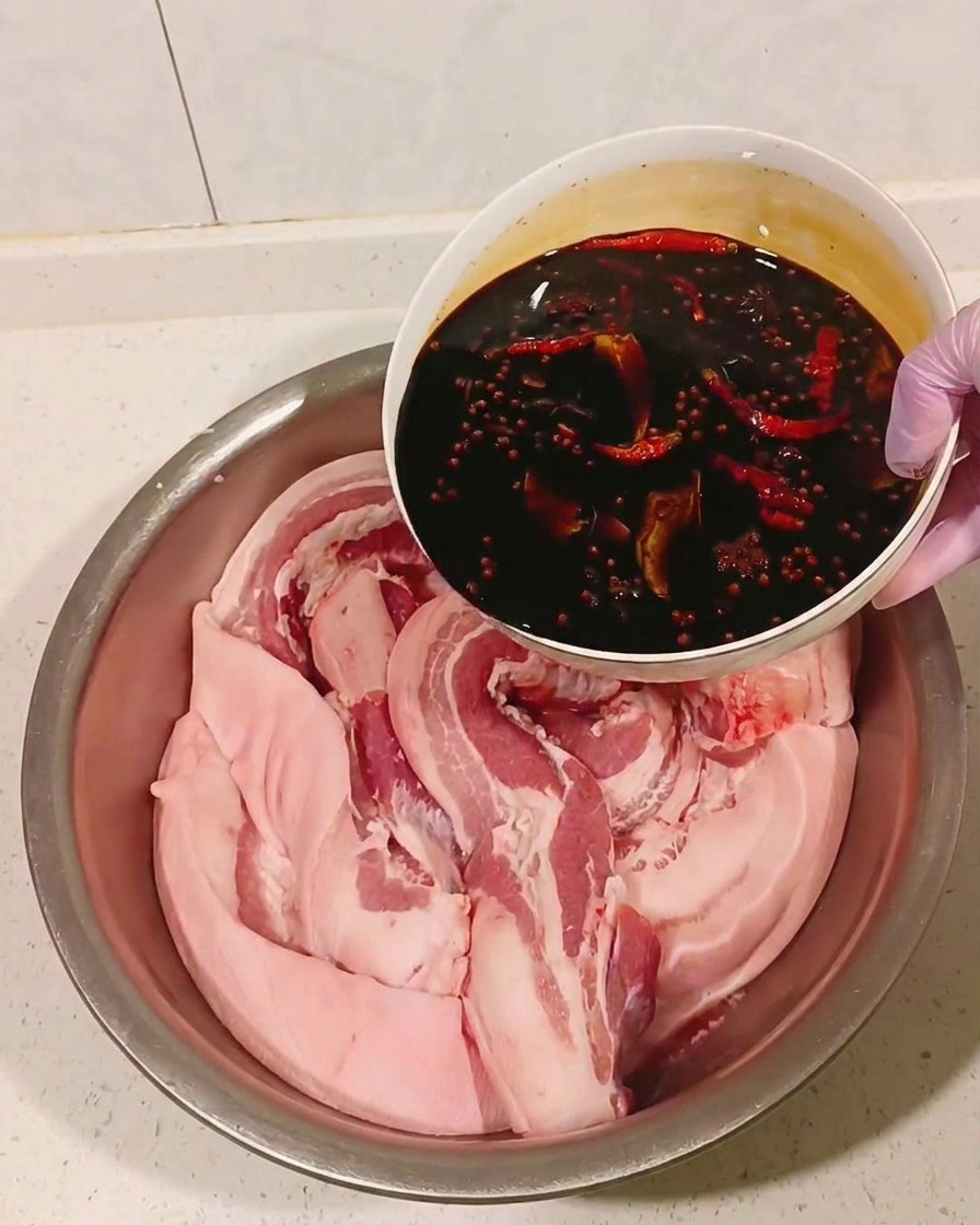 Abgekühlte, würzige Sojasaucen-Mischung wird über den Schweinebauch in einer Metallschüssel gegossen.