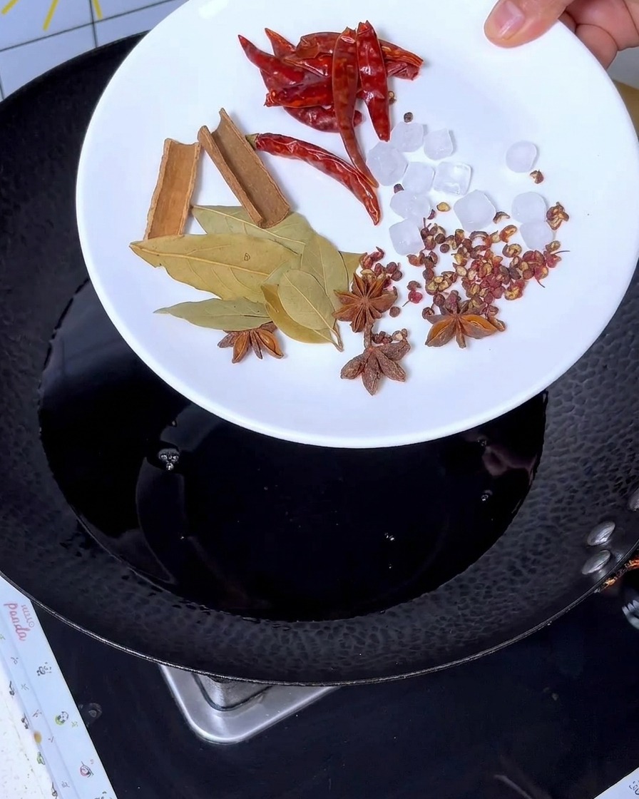 Gewürze wie Zimt, Sternanis, Lorbeerblätter, Chili und Kandiszucker werden zur dunklen Flüssigkeit im Wok gegeben.