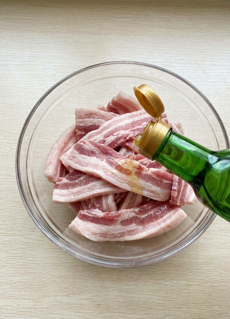 Sojasauce wird aus einer Glasflasche über rohe Schweinebauchstreifen in einer Schüssel gegossen.