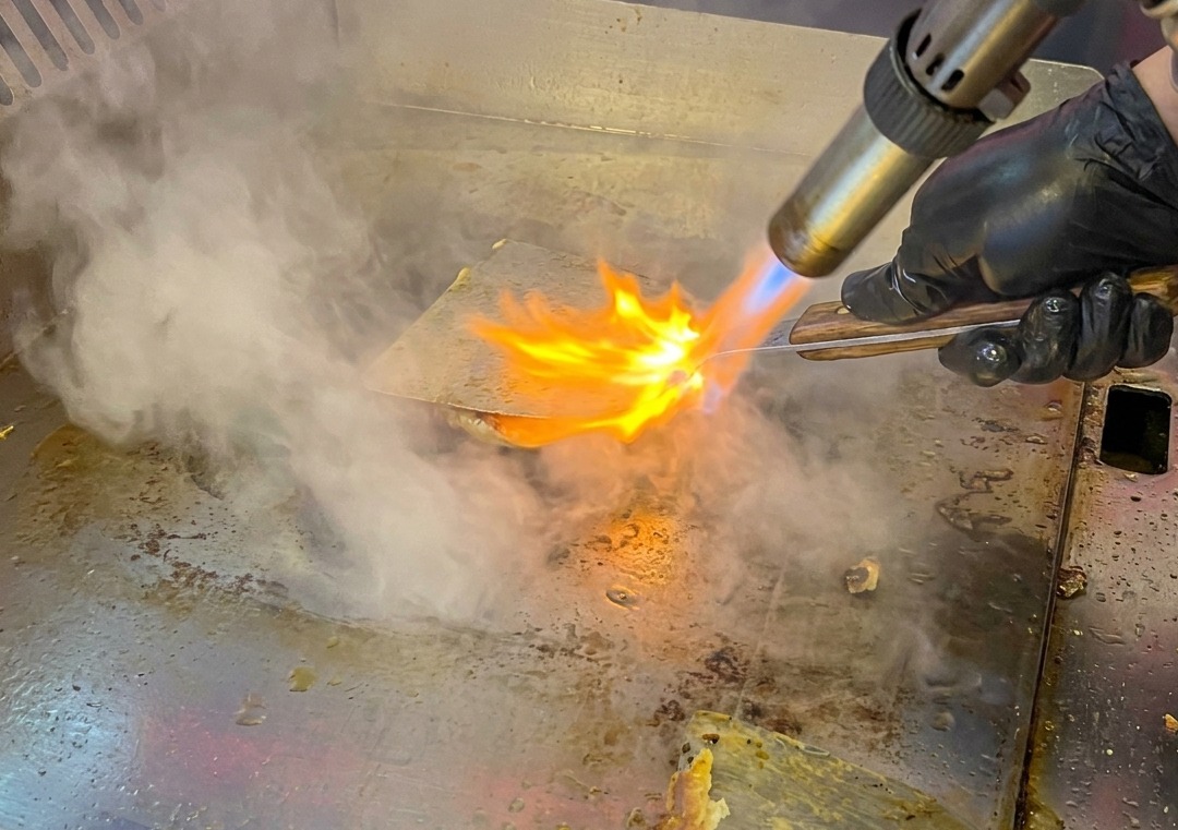 Ein Rinder-Patty auf der Grillplatte wird mit einem Küchenbrenner flambiert.