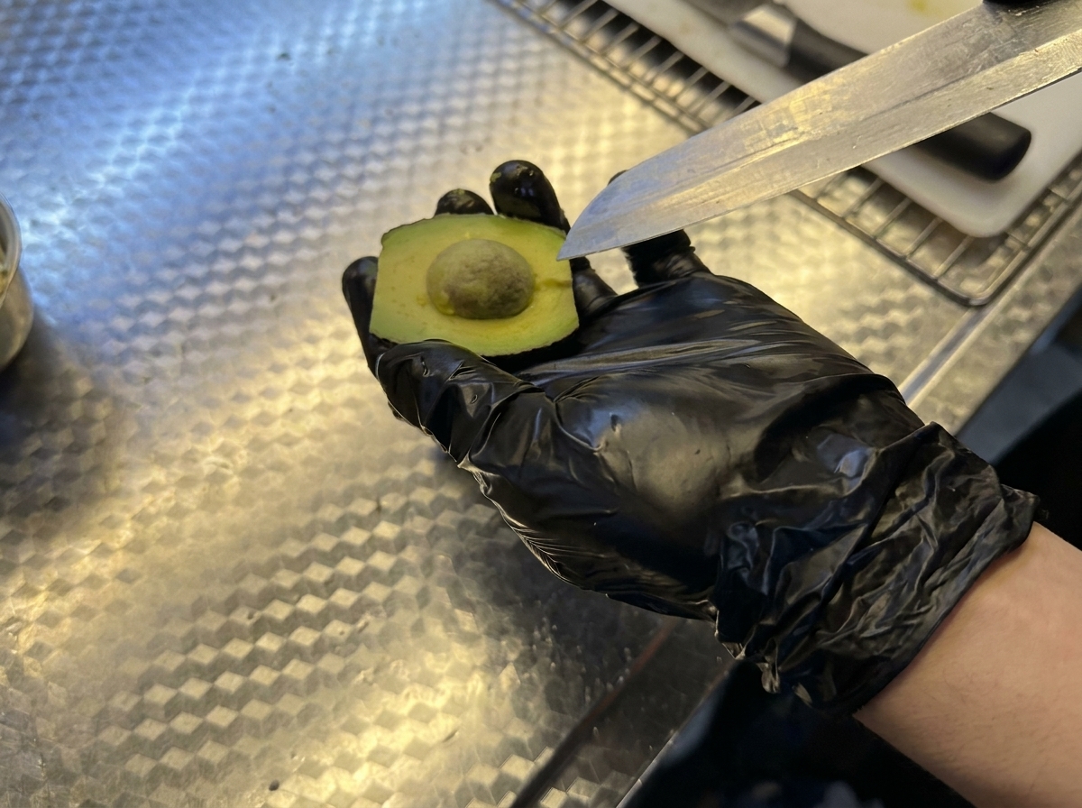 Hand mit schwarzem Handschuh hält eine Avocadohälfte, während der Kern mit einem Messer entfernt wird.