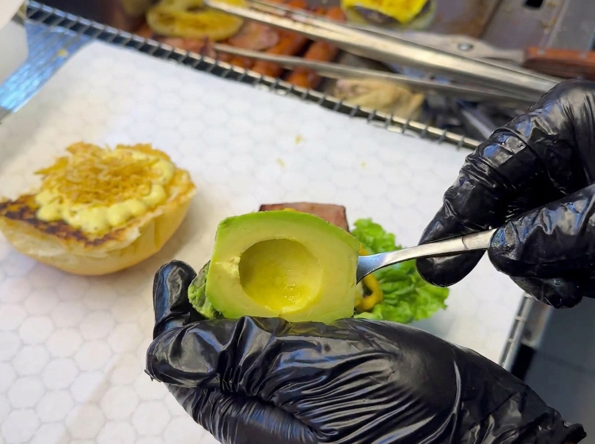 Hand mit schwarzem Handschuh platziert Avocadoscheiben auf die bereits belegte Burgerbasis.