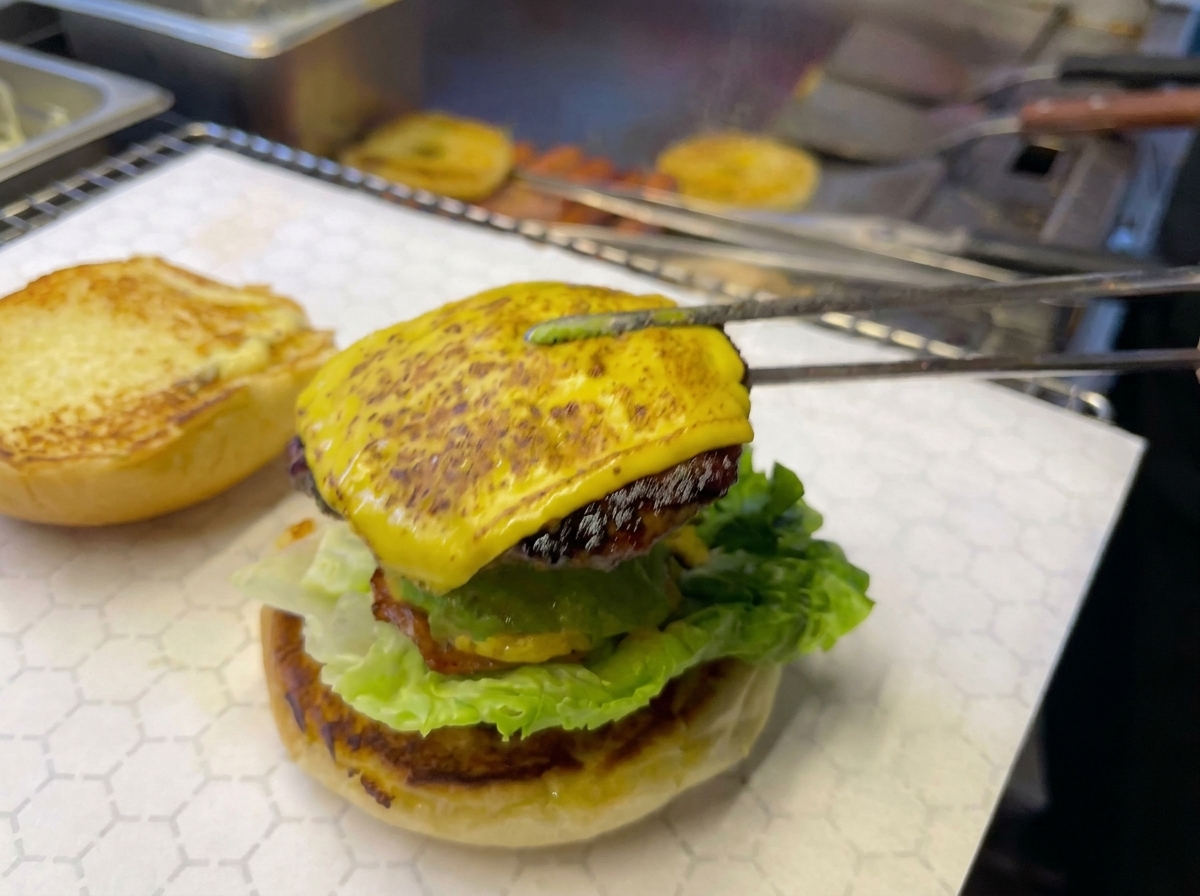 Eine Grillzange platziert das Käse-Patty mit Ananasscheibe auf den Burgeraufbau.