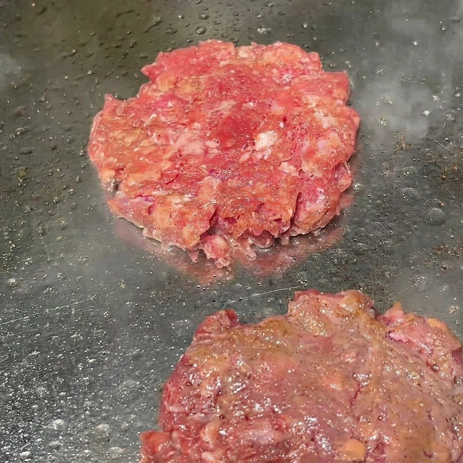 Rindfleisch-Patties auf dem Grill werden mit frisch gemahlenem schwarzen Pfeffer bestreut.