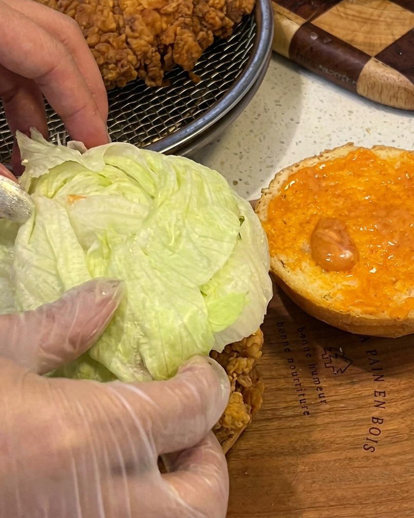 Ein Burger wird mit knusprigem Filet, frischem Salat und Sauce auf einem gerösteten Brötchen zusammengebaut.