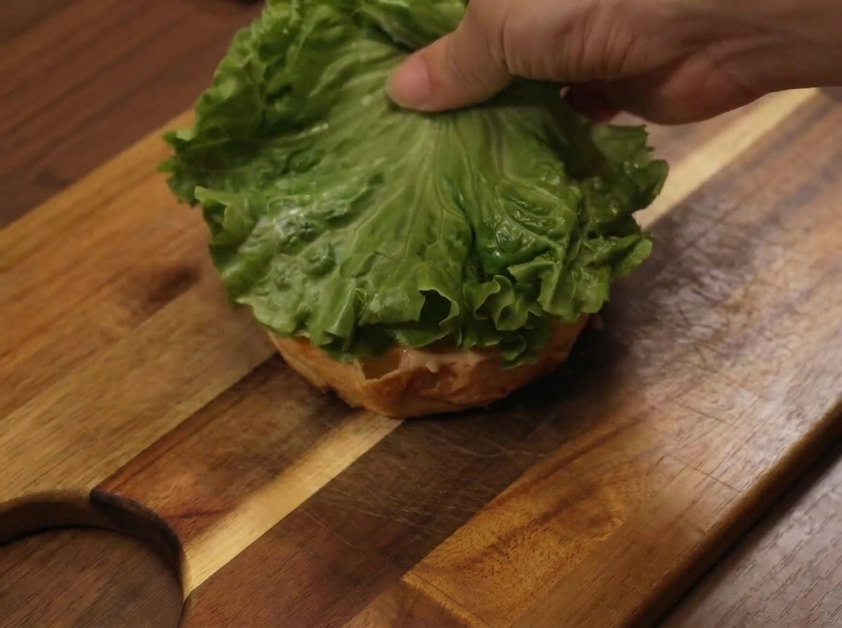 Ein Salatblatt wird beim Zusammensetzen des Burgers auf die untere Brötchenhälfte gelegt.
