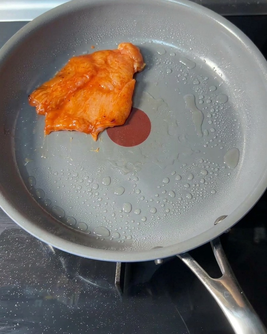 Rohe, marinierte Hähnchen-Patties liegen in einer Pfanne mit etwas Speiseöl.