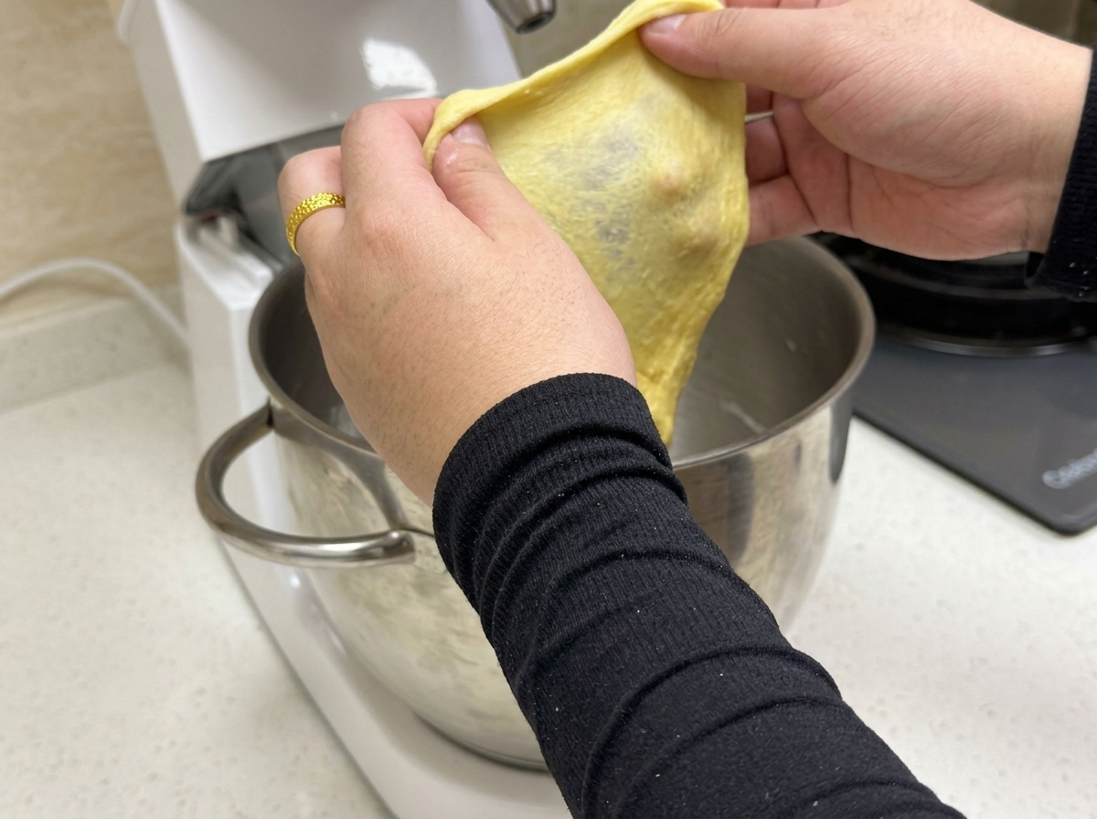 Ein kleines Stück gelber Teig wird gedehnt, um eine dünne, elastische Glutenmembran zu zeigen.