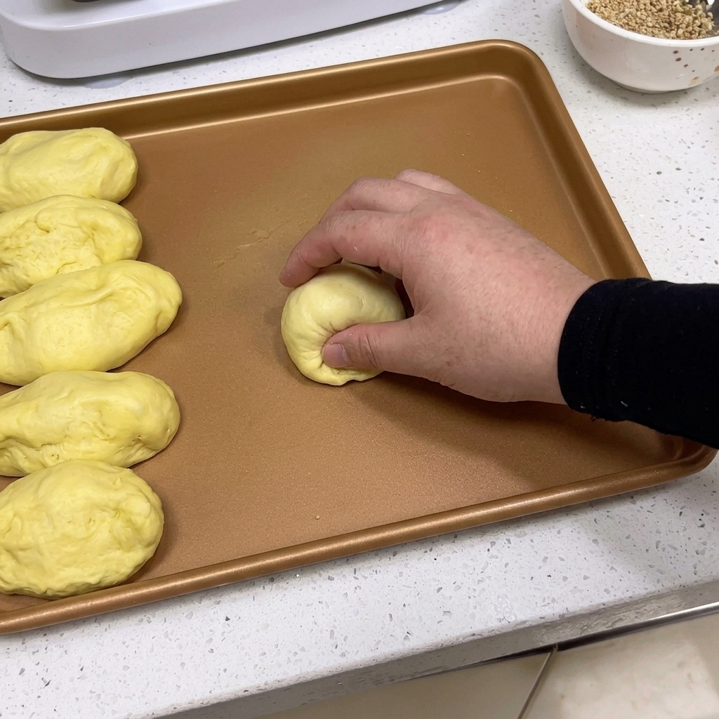 Hände formen eine Teigportion zu einer glatten, runden Kugel auf einem Backblech.