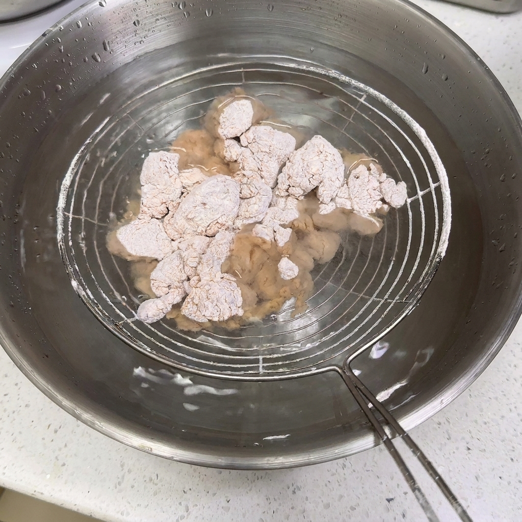 Mit Mehl bestäubte Hähnchenstücke werden auf einem Schaumlöffel in eine Edelstahlschale mit Wasser getaucht.