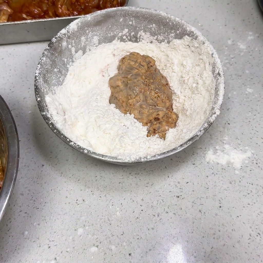 Ein Stück nasses, paniertes Hähnchen wird in einer Silberschüssel mit trockenem Mehl gewendet.