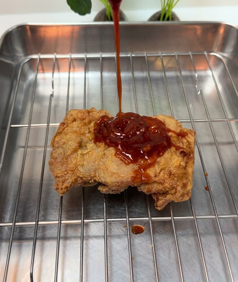 Dicke rote Sauce wird über ein knuspriges Hähnchenstück auf einem Metallgitter gegossen.