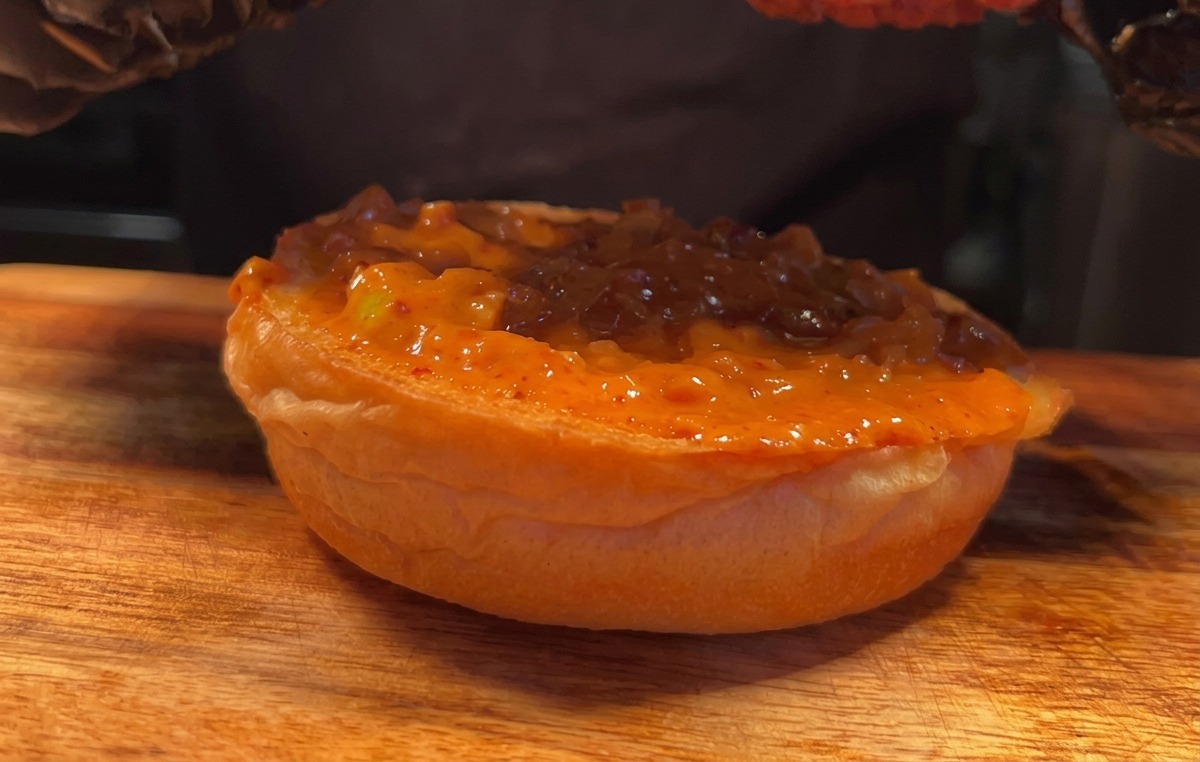 Ein mit Handschuh bekleideter Hand platziert ein knuspriges Hähnchenstück auf dem Brötchen.