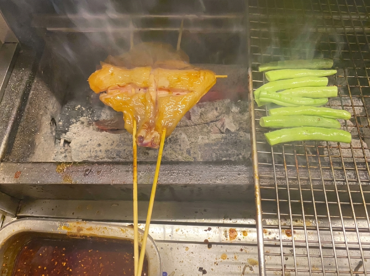 Gespießtes Hähnchen und grüne Bohnen garen auf einem Metallgrill über Holzkohle.