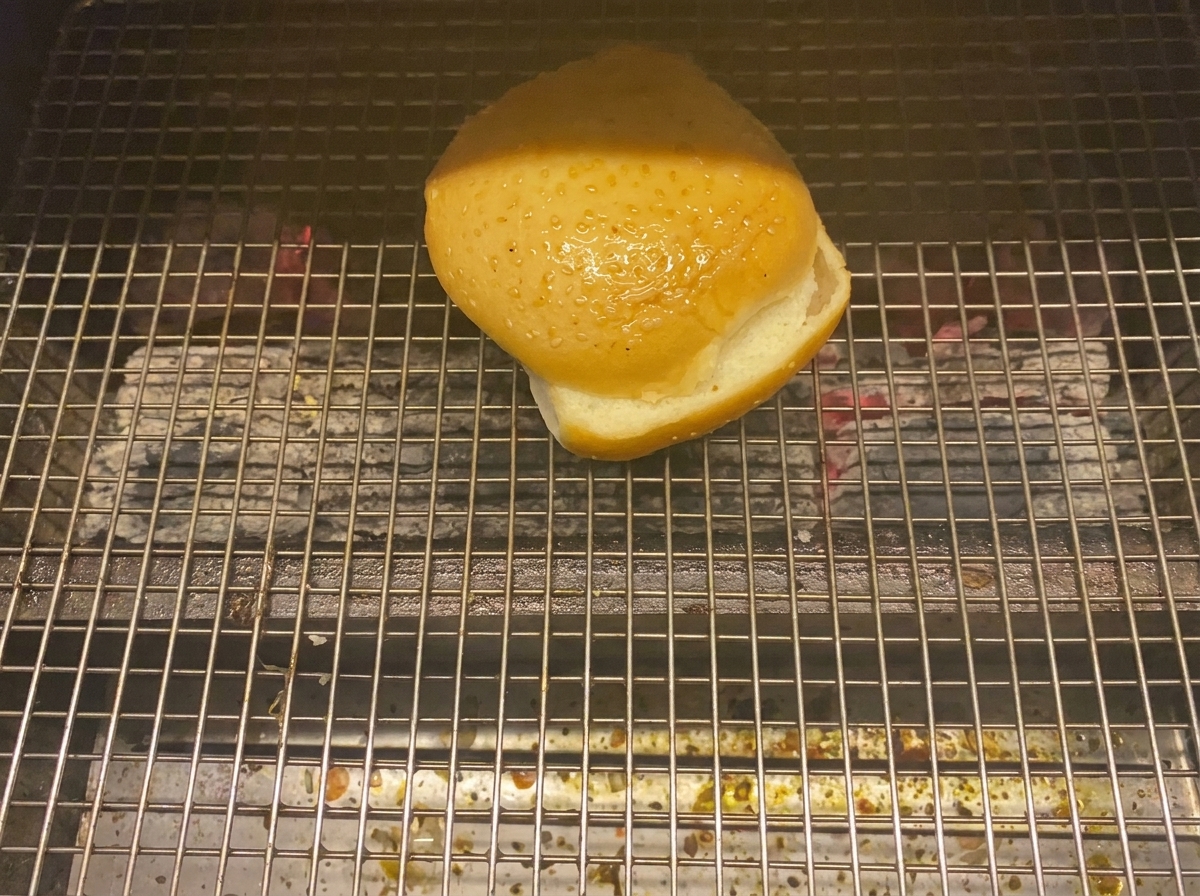 Eine Burgerbrötchenhälfte röstet mit der Schnittfläche nach unten auf einem Holzkohlegrill.