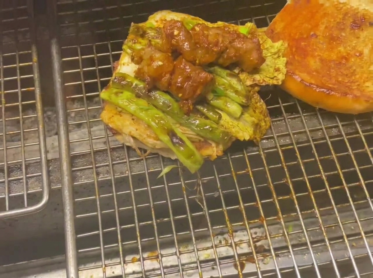 Die untere Hälfte eines Burgerbrötchens, belegt mit gegrilltem Gemüse und glasierten Fleischspießen auf einem Grill.