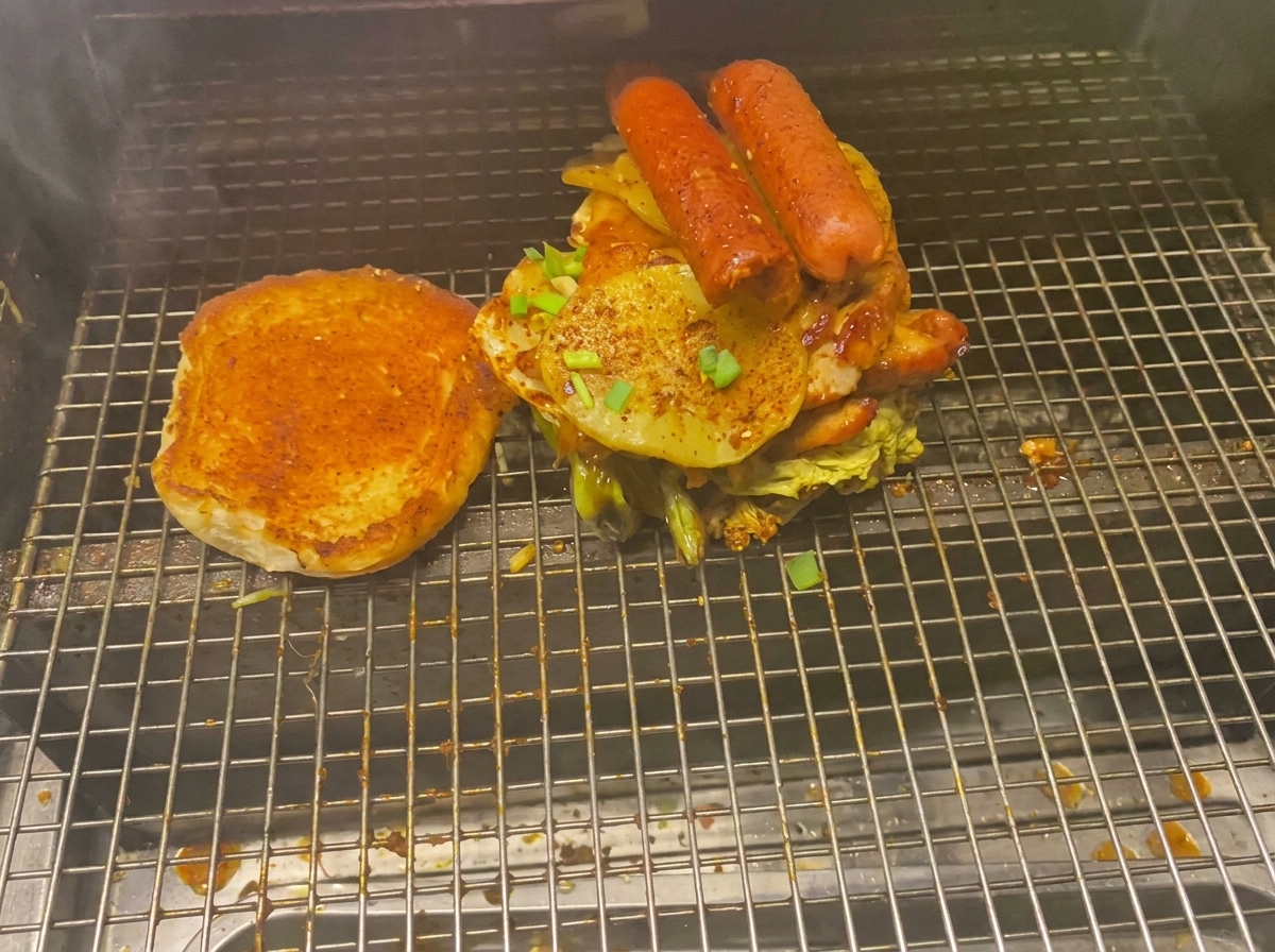 Ein turmhoher Burger, beladen mit Fleisch, gegrillten Kartoffeln und Würsten neben einem gerösteten Brötchen auf einem Grillrost.