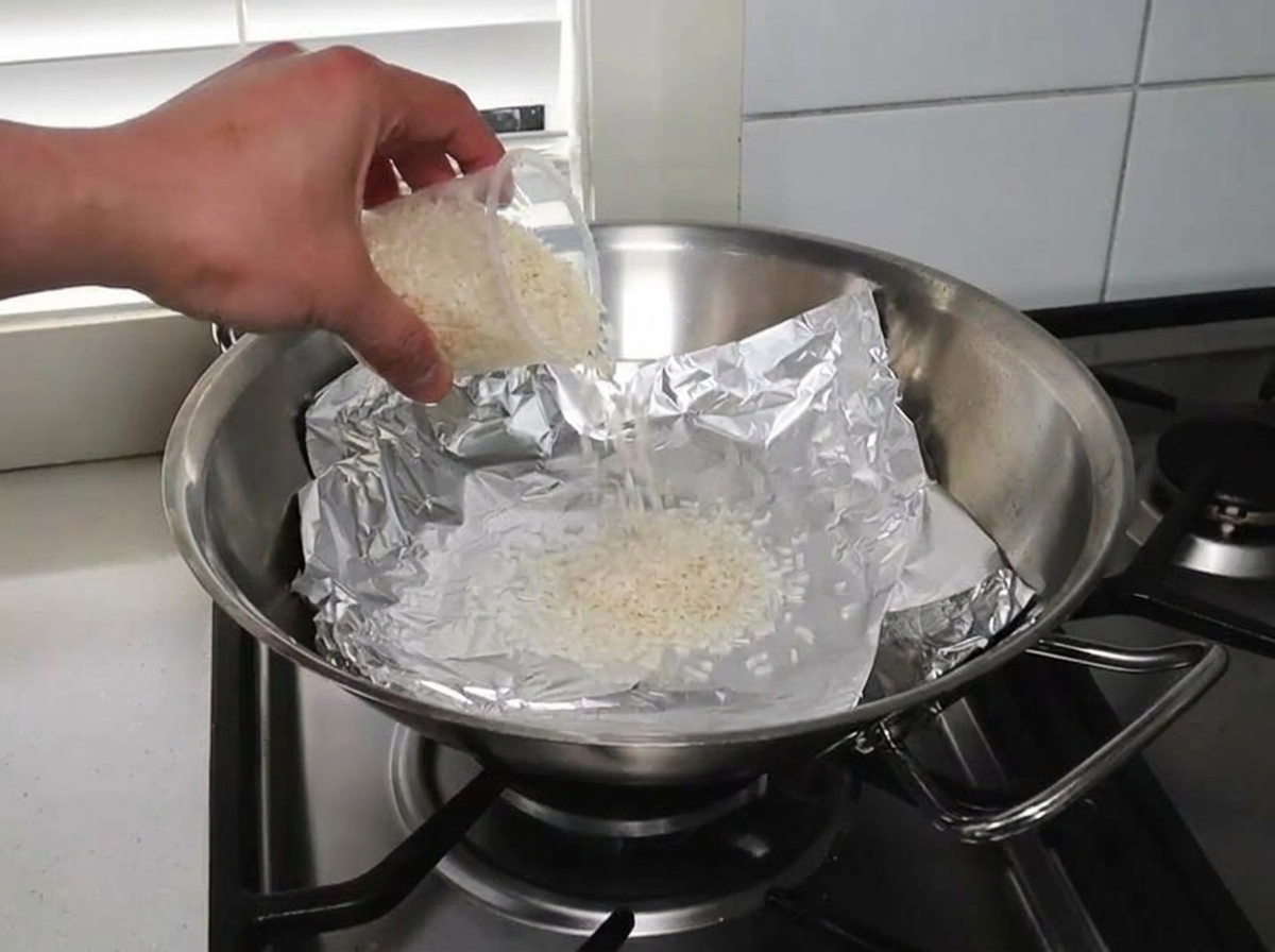 Roher Reis und Räucherspäne sind gleichmäßig auf einem mit Alufolie ausgelegten Wok verteilt.