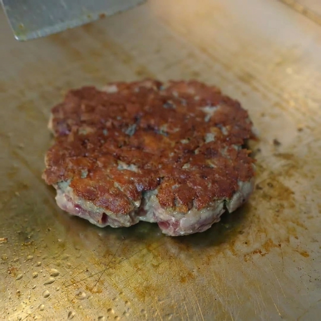 Eine Scheibe gelber Käse, die auf einem gebratenen Rindfleisch-Patty unter einer Metallhaube schmilzt.
