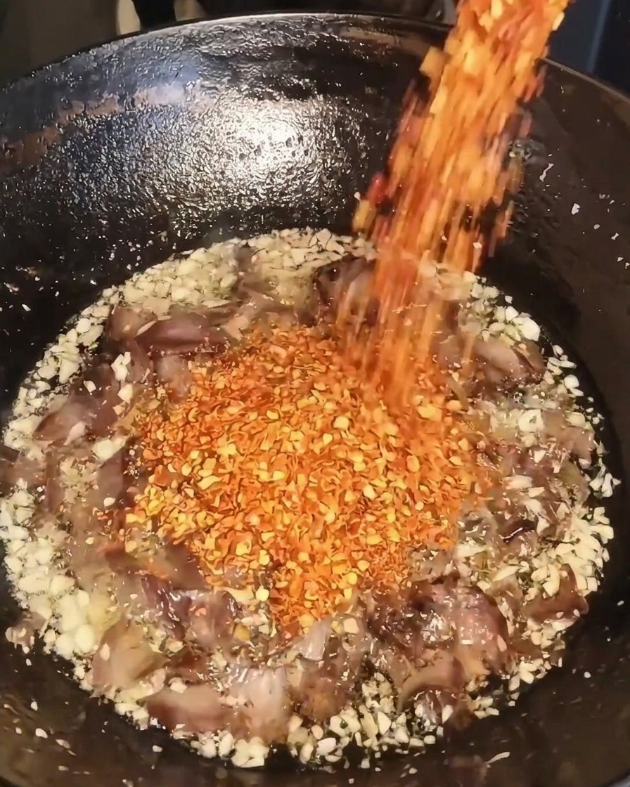 Getrocknete rote Chiliflocken werden aus einem Behälter in einen Wok mit gebratenem Knoblauch, Rindfleisch und Öl geschüttet.