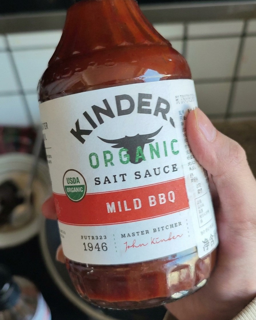 Eine Hand hält eine Flasche mit milder BBQ-Sauce und bereitet sich darauf vor, sie über das Fleisch zu geben.