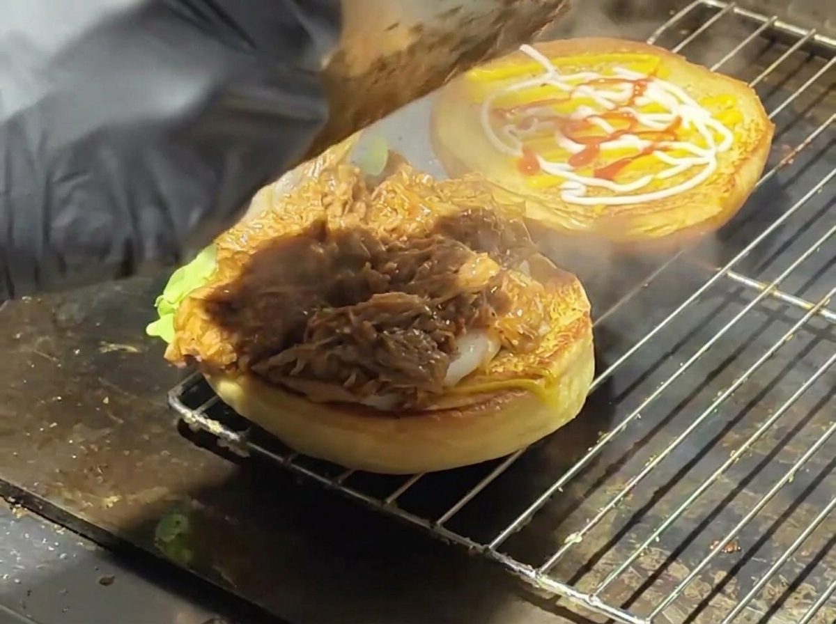 Saftiges Pulled Pork wird auf einem getoasteten Burgerbrötchen mit Salat verteilt.