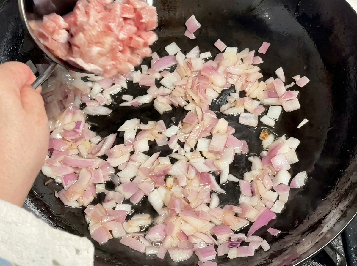 Rohes gewürfeltes Schweinefleisch wird aus einer Metallschüssel in einen Wok mit angebratenen roten Zwiebeln gegeben.