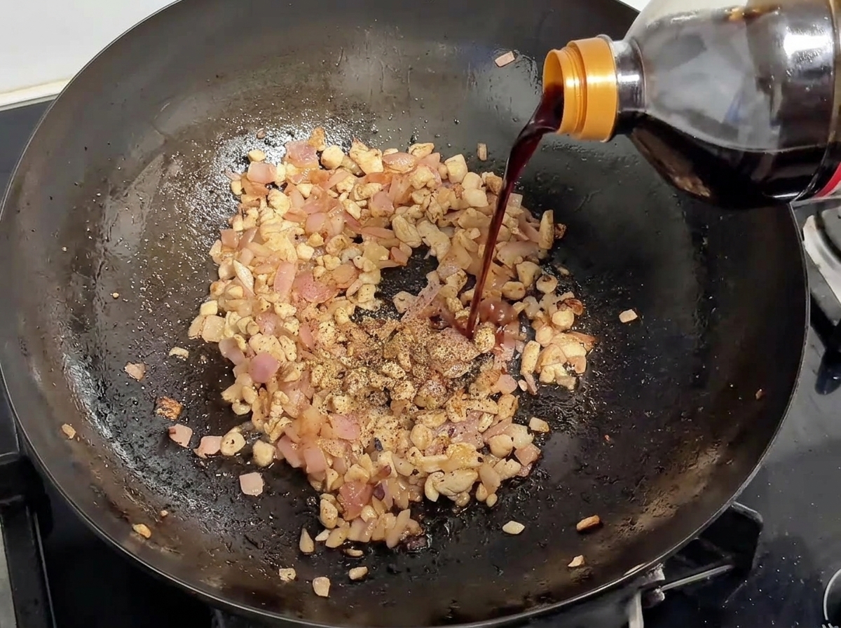 Sojasauce wird aus einer Flasche in einen Wok mit gebräuntem Schweinefleisch und Zwiebeln gegossen, garniert mit schwarzem Pfeffer.