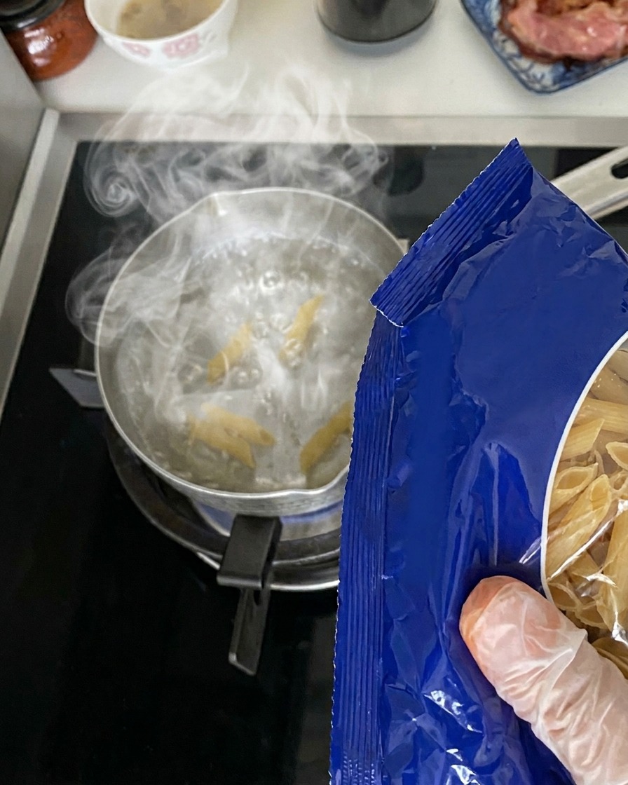 Trockene Penne-Nudeln werden aus einer blauen Verpackung in einen Metalltopf mit kochendem Wasser auf dem Herd gegossen.