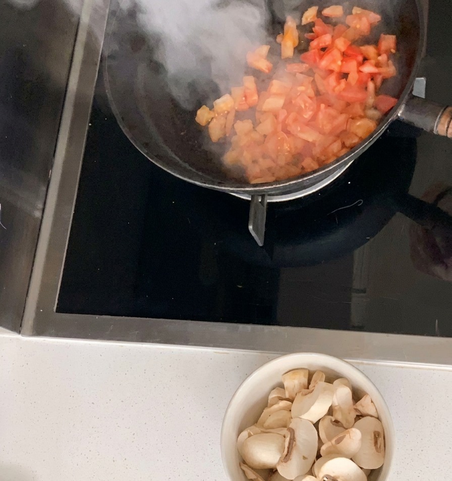 Gewürfelte Tomaten werden in einer schwarzen Pfanne angebraten, während eine Schüssel mit in Scheiben geschnittenen weißen Champignons darunter auf der Arbeitsfläche steht.