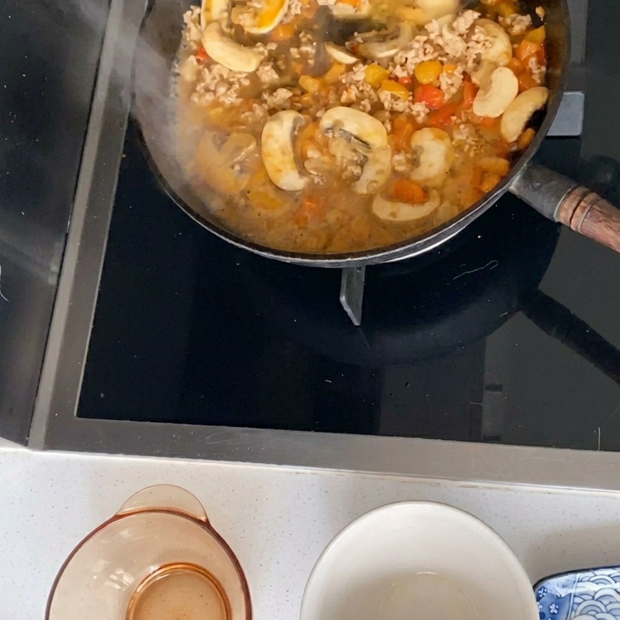Eine köchelnde Sauce aus gehacktem Schweinefleisch, in Scheiben geschnittenen Champignons und gewürfelten Tomaten blubbert in einer schwarzen Bratpfanne.