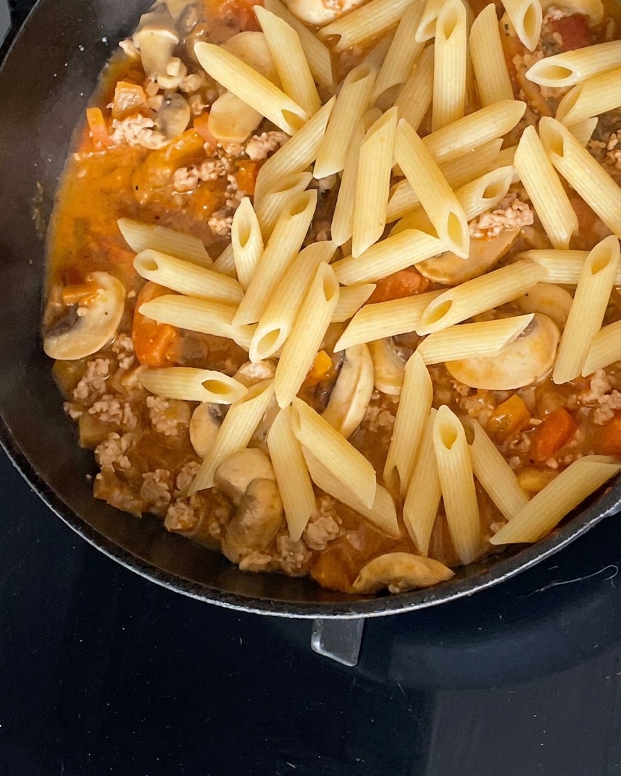 Gekochte Penne-Nudeln ruhen auf einer herzhaften Fleisch-, Pilz- und Tomatensauce in einer schwarzen Pfanne.