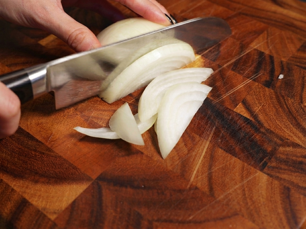 Eine Hand schneidet mit einem Küchenmesser eine Zwiebel auf einem Holzbrett.