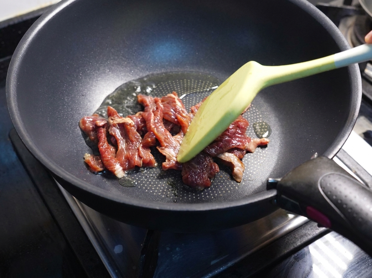 Mit einem Pfannenwender wird Fleisch in einer dunklen Pfanne angebraten.