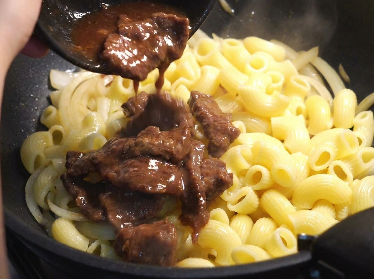 Angebratenes Rindfleisch wird zu den Makkaroni und Zwiebeln in die Pfanne gegeben.