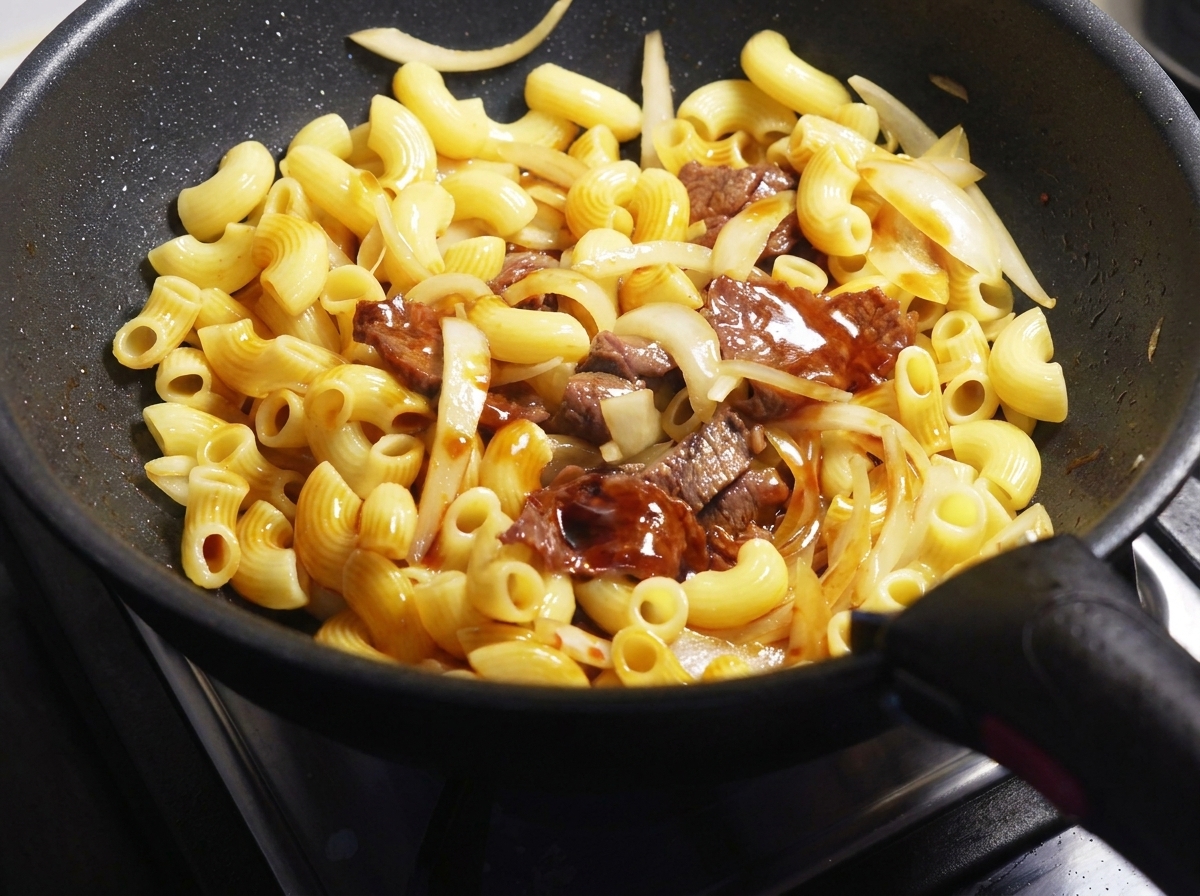 Dunkle Sojasauce wird über Nudeln, Fleisch und Zwiebeln in einer schwarzen Pfanne gegossen.