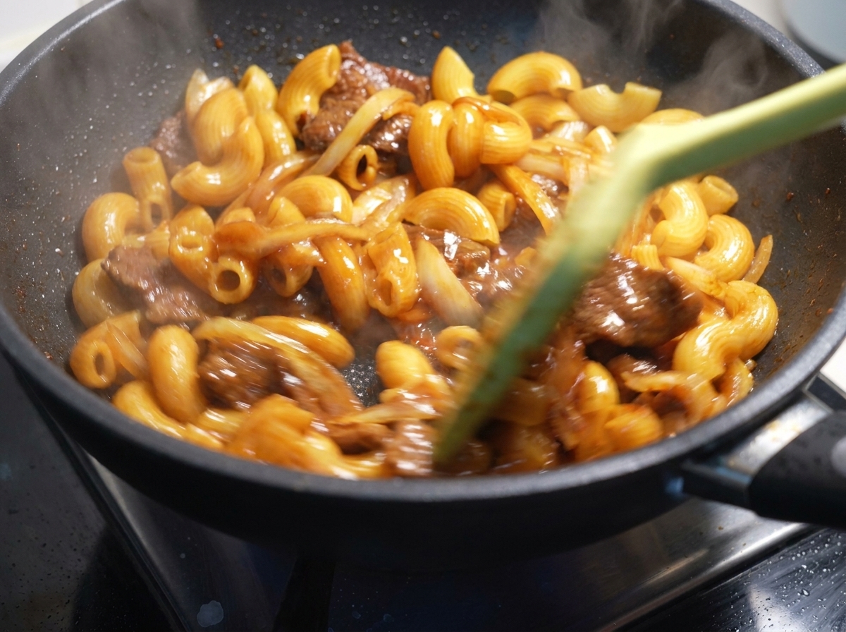 Makkaroni, Fleisch und Zwiebeln werden in einer heißen Pfanne kräftig durchgeschwenkt.