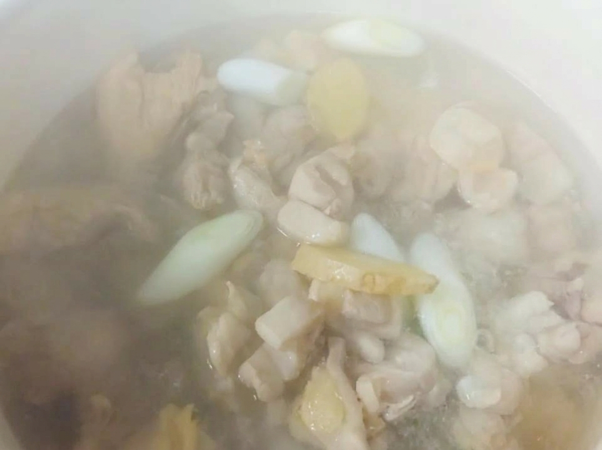 Heißes Wasser dampft in einem weißen Topf über Hähnchenteilen, geschnittenem Ingwer und Frühlingszwiebeln.