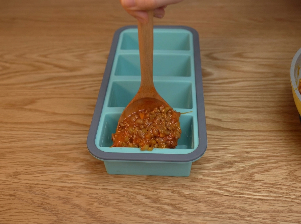 Dicke Tomaten-Hackfleisch-Sauce wird in einzelne Fächer einer blauen Silikonform gelöffelt.