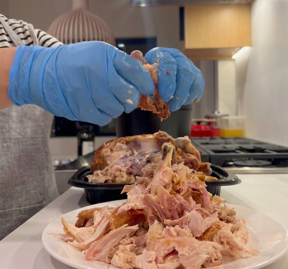 Hände in blauen Küchenhandschuhen zupfen das Fleisch eines Brathähnchens auf einen weißen Teller.