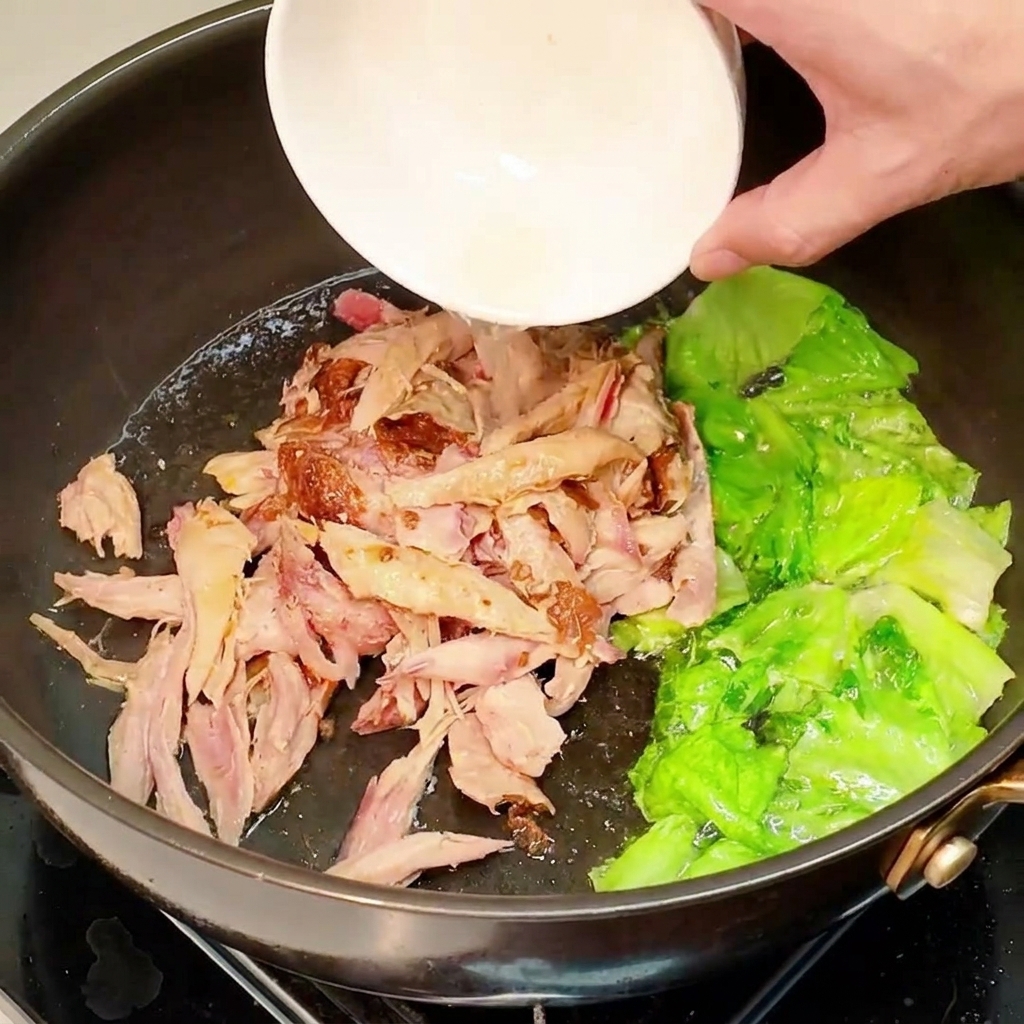 Vorgegartes, zerkleinertes Hähnchen wird aus einer kleinen weißen Schüssel in die Pfanne mit angebratenem Salat gegeben.